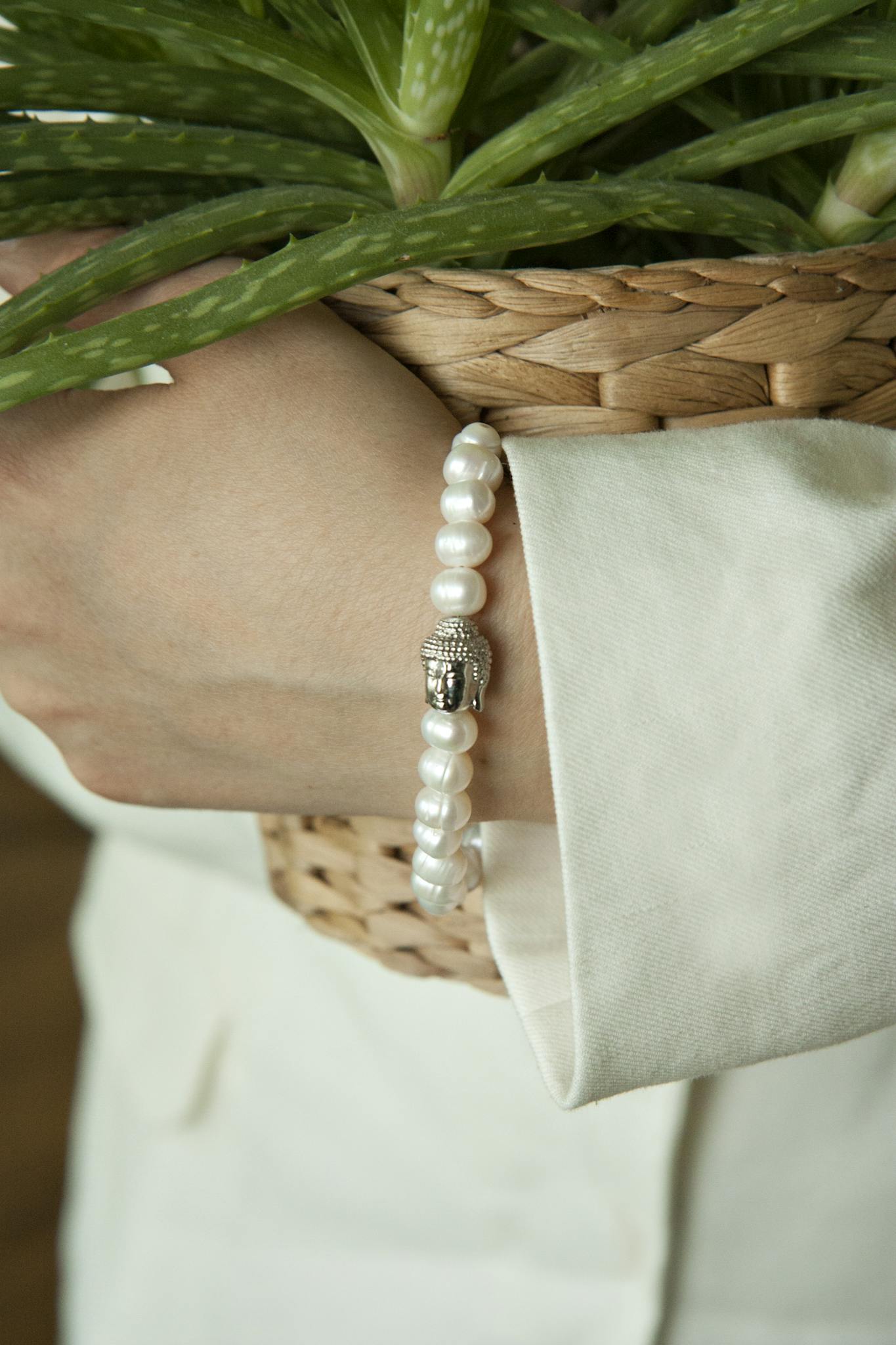 Close-up of a hand wearing a pearl bracelet with a Buddha charm, holding a basket with green plants.